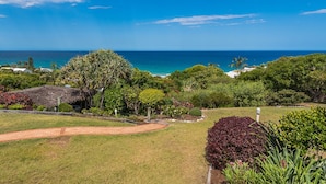A lush garden with a variety of plants and trees, a thatched-roof structure, and a view of the ocean. - Whale season is upon us, watch from your balcony (SUNSHINE BEACH)