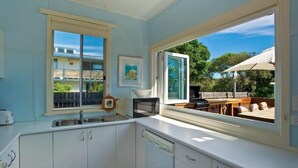 A modern kitchen with white cabinets, a built-in microwave, and a large window offering a view of the outdoor patio area. - Two Bedroom House Wooli (Wooli)