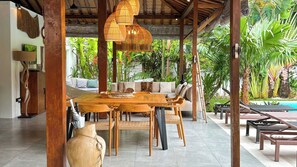 Een overdekt terras met een houten tafel en stoelen, omgeven door weelderige tropische planten en een zwembad.