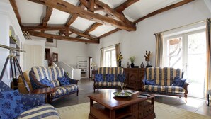 Un salón espacioso con vigas de madera, dos sofás estampados, una mesa de centro y un gran ventanal con cortinas.