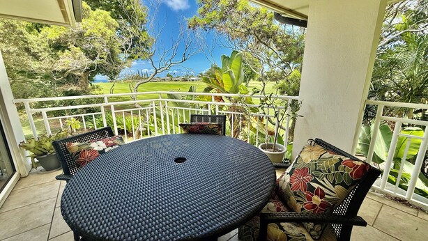 A round table with a woven tabletop and two chairs with floral upholstery, set on a balcony with a white railing and potted plants.