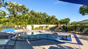 A swimming pool with lounge chairs and a covered area, surrounded by palm trees and a white wall with plants.