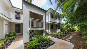 A two-story building with a balcony on the second floor, surrounded by lush greenery and a garden path.