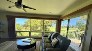 A living room with a leather sofa, a coffee table, and a ceiling fan. Large windows offer a view of a green landscape.