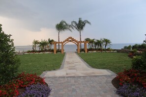 A pathway leading to a wooden archway with palm trees and flower beds on either side.