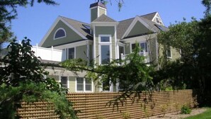 A large, two-story house with a prominent chimney, multiple windows, and a wooden fence in the foreground. - Expansive Home With Gorgeous Bay Views! (1052) (Brewster)