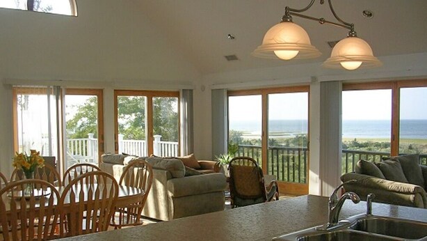 A spacious kitchen with a large island, a dining area with wooden chairs, and large windows offering a view of the ocean. - Expansive Home With Gorgeous Bay Views! (1052) (Brewster)