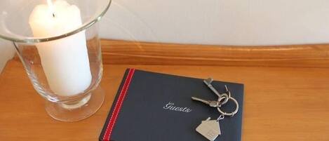 Een kaars in een glazen houder, een zwart notitieboekje met een rode lint, en een sleutelhanger met een huisje aan.