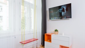 Et moderne værelse med fjernsyn ophængt på væggen, en rød bænk, en hvid kommode og et vindue med lette gardiner.