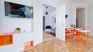 Et moderne værelse med et fjernsyn, der viser en film, et spiseområde med orange stole og en seng med et stribet tæppe.