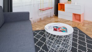 En moderne stue med en grå sofa, et rundt sofabord, et fjernsyn monteret på væggen og et vindue med lette gardiner.