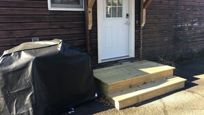 Una cabaña de madera con una puerta blanca, un pequeño porche con escalones y un objeto cubierto en el suelo.