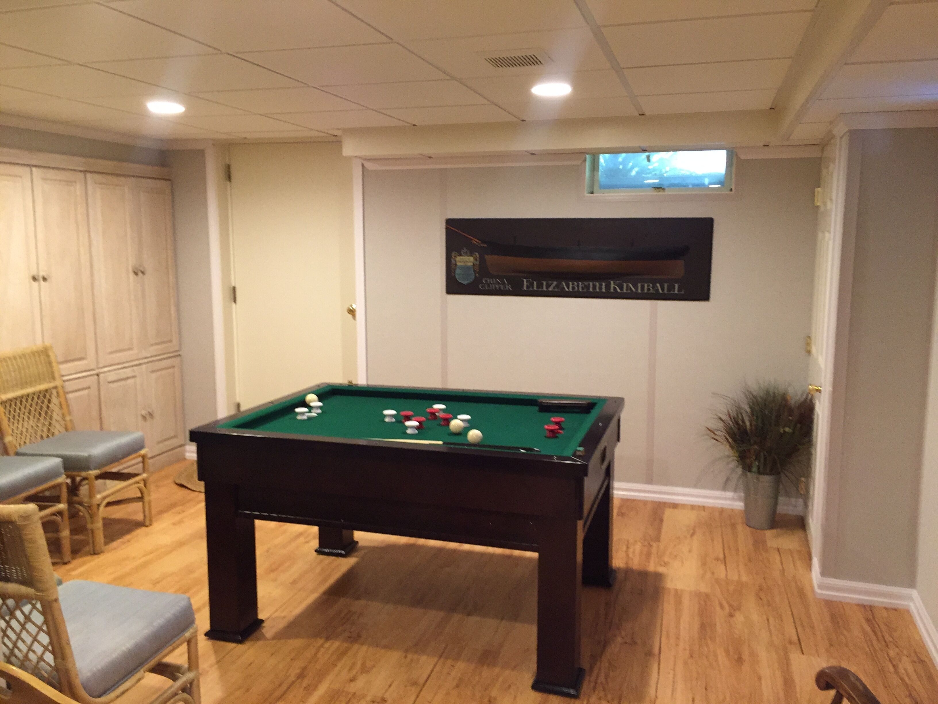 A pool table with balls, a chair, and a potted plant in a room with wooden flooring and a ceiling light.