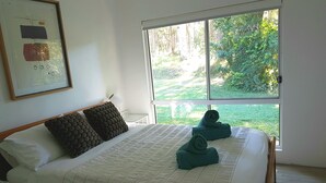 Een netjes opgemaakt bed met een witte deken, twee donkere kussens en twee opgerolde handdoeken. Aan de muur hangt een lijst met abstracte kunst, een raam met uitzicht op groen en een witte nachtkastje.