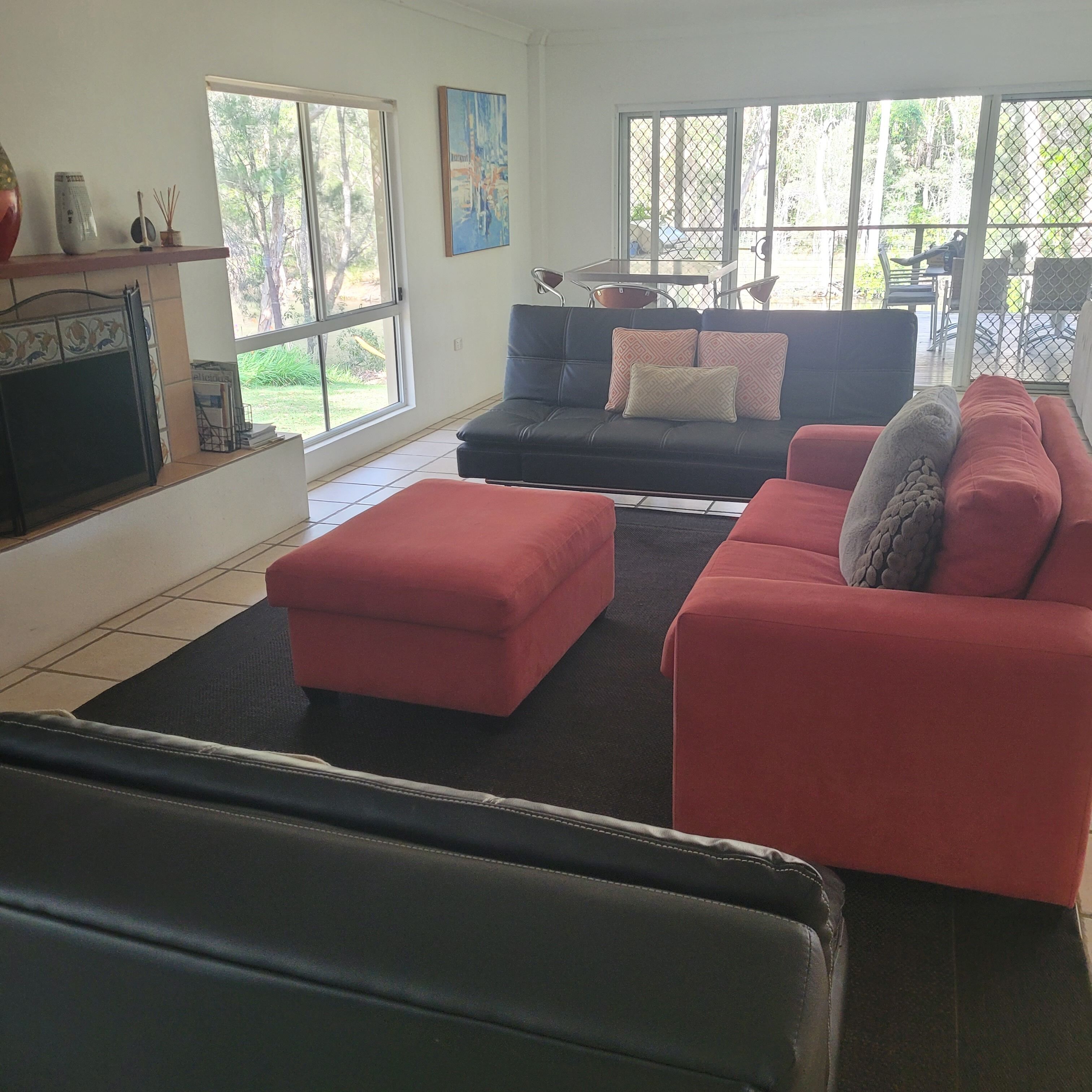 A living room with a black sofa, red ottoman, and a black leather chair. There is a TV on the left and a window with a view of greenery outside.