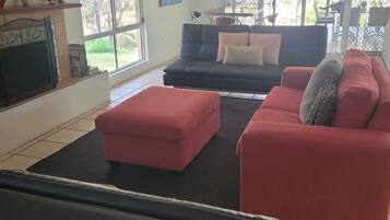 A living room with a black sofa, red ottoman, and a black leather chair. There is a TV on the left and a window with a view of greenery outside.