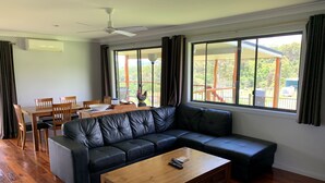 A modern living room with a sectional sofa, a wooden coffee table, and a dining area with a table and chairs. Large windows offer a view of the outdoors.