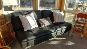 Un canapé en cuir noir, agrémenté de coussins à rayures et à pois, est placé sur une terrasse en bois, d’où l’on aperçoit une clôture en bois et une voiture au loin.