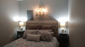 Une chambre à coucher avec une tête de lit capitonnée, une literie à motifs, deux lampes de chevet et un luminaire fixé au mur.