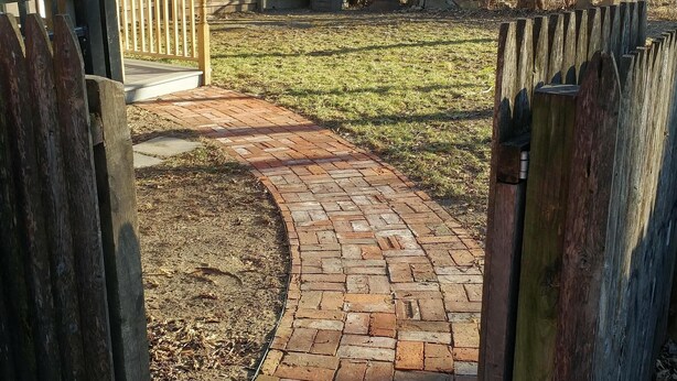 Un chemin pavé de briques mène à une zone délimitée par une barrière et entourée d’une clôture en bois.