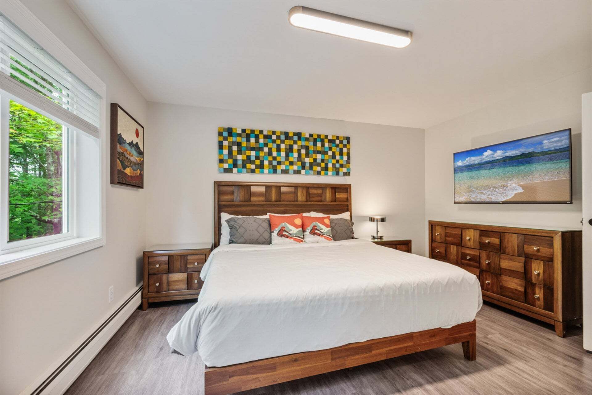 Une chambre à coucher avec un lit en bois, une table de chevet, une commode, un tableau et une fenêtre donnant sur un paysage verdoyant.