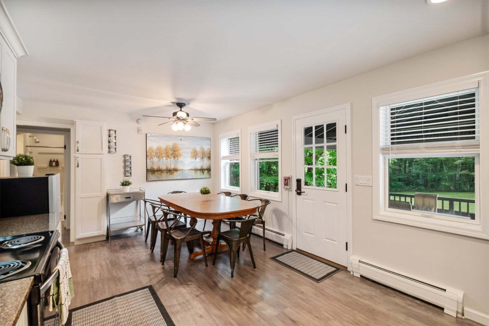 A kitchen with a dining table, chairs, a ceiling fan, and a view of greenery through the windows.