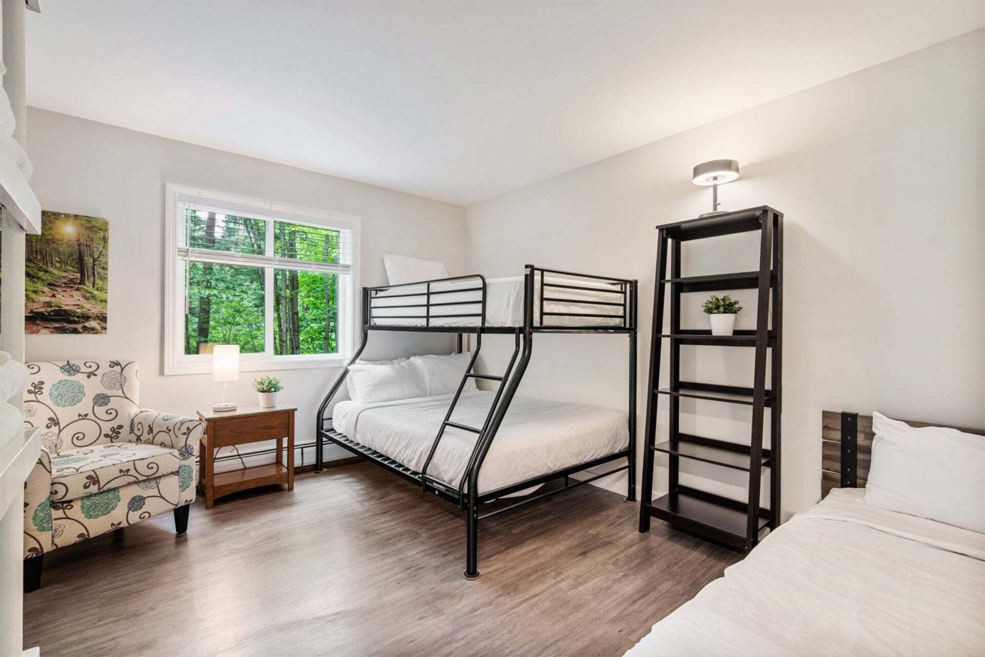 A bunk bed room with a window, a bedside table with a lamp, a patterned chair, and a shelving unit.