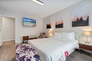 Hotellihuone, jossa on suuri sänky, yöpöytä lampun kanssa, tuoli ja taideteoksia seinillä.