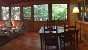 A wooden deck with a table and chairs, large windows with a forest view, and a lamp on the right. - Boathouse Cottage. Private Beach. Sunset, Mountain,  Lake Views. Near Acadia NP! (Sullivan)