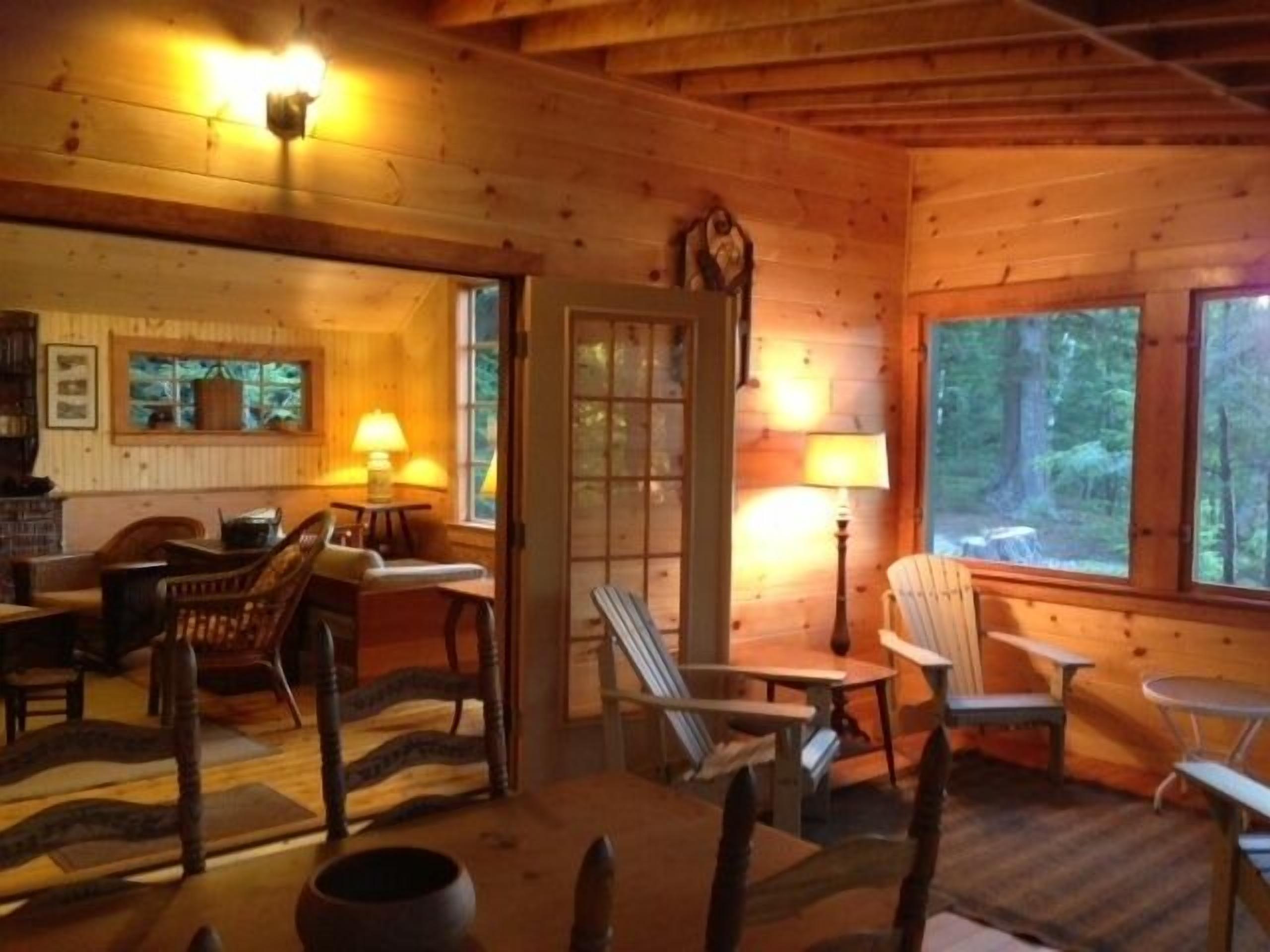 A cozy cabin interior with wooden walls, a fireplace, a dining area with chairs, and large windows offering a view of the forest.