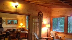 A cozy cabin interior with wooden walls, a fireplace, a dining area with chairs, and large windows offering a view of the forest. - Boathouse Cottage. Private Beach. Sunset, Mountain,  Lake Views. Near Acadia NP! (Sullivan)