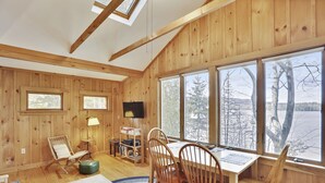 L’intérieur chaleureux d’un chalet en bois comprend un coin repas, un téléviseur et une grande fenêtre donnant sur un paysage enneigé.