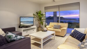 Un salon moderne doté d’un téléviseur à écran plat, d’un canapé, d’une table basse et d’une grande fenêtre offrant une vue sur la plage au coucher du soleil.