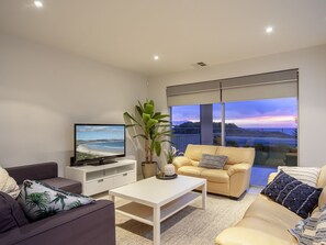 Ein modernes Wohnzimmer mit einem Flachbildfernseher, einer Couch, einem Couchtisch und einem großen Fenster mit Blick auf den Strand bei Sonnenuntergang.