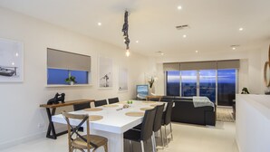 Un espace de salle à manger moderne, avec une table blanche, des chaises noires et une vue sur l’océan grâce à de grandes fenêtres.