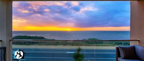A balcony with a view of the sunset over the ocean, a glass door, a small table with a vase, and a sofa.