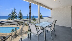 Un balcón con una mesa y sillas de vidrio, con vista a una piscina y una playa, árboles y un cielo despejado.