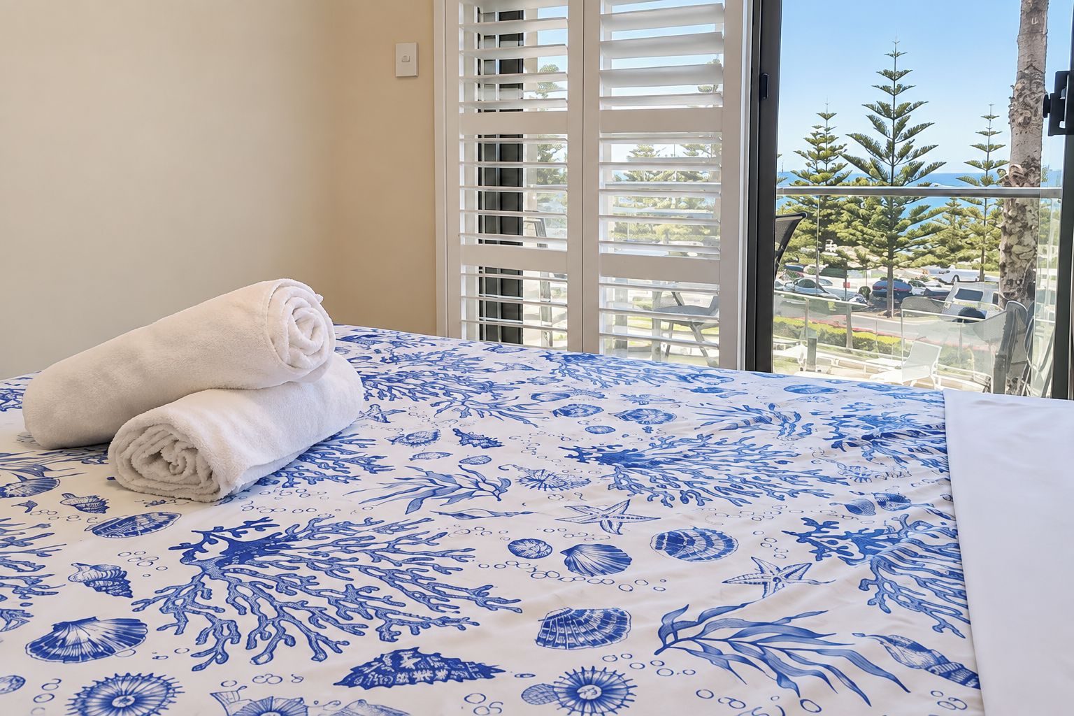 A bed with a floral bedspread, a pair of rolled towels, and a view of a balcony with trees and parked cars.
