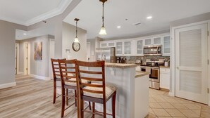 A modern kitchen with white cabinets, stainless steel appliances, and a central island. There are two wooden chairs with cushioned seats. - OVER-SIZED 2 BR PATIO UNIT STEPS TO POOL & COURTYARD!!! (Henderson)