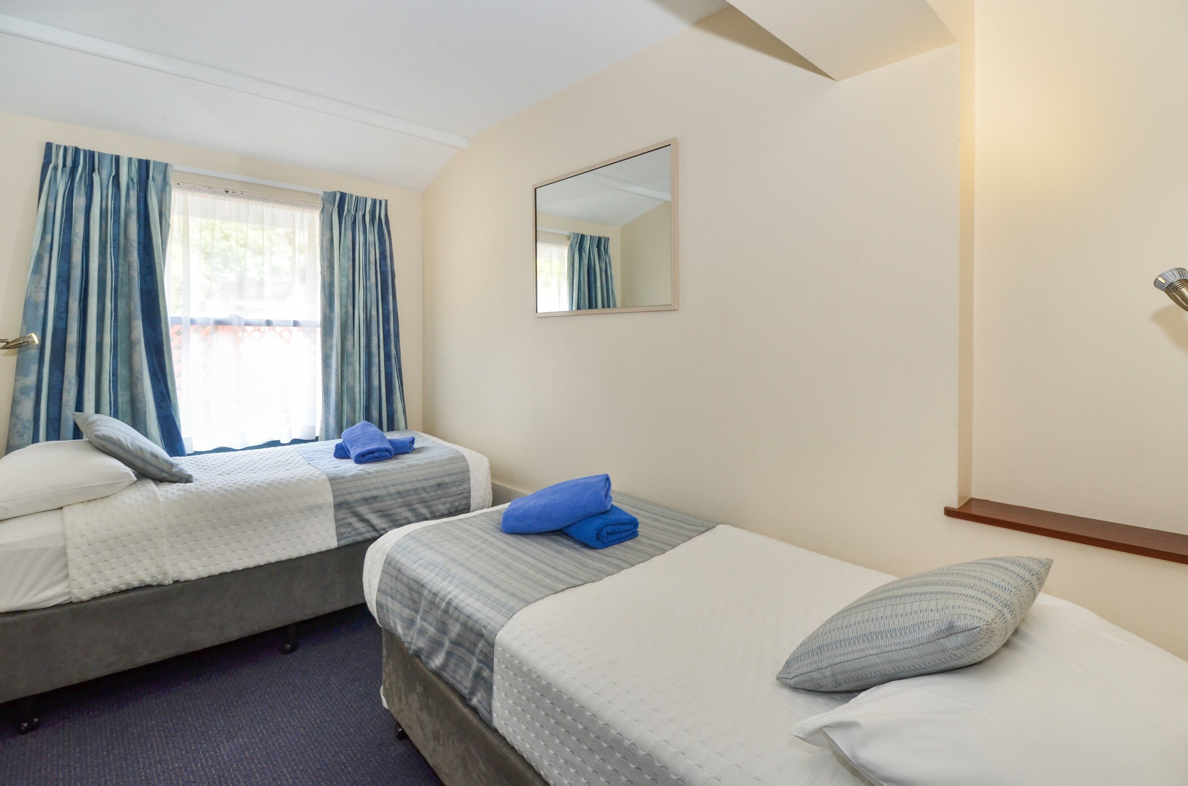 A hotel room with two beds, a window with curtains, a mirror, and a wooden shelf.