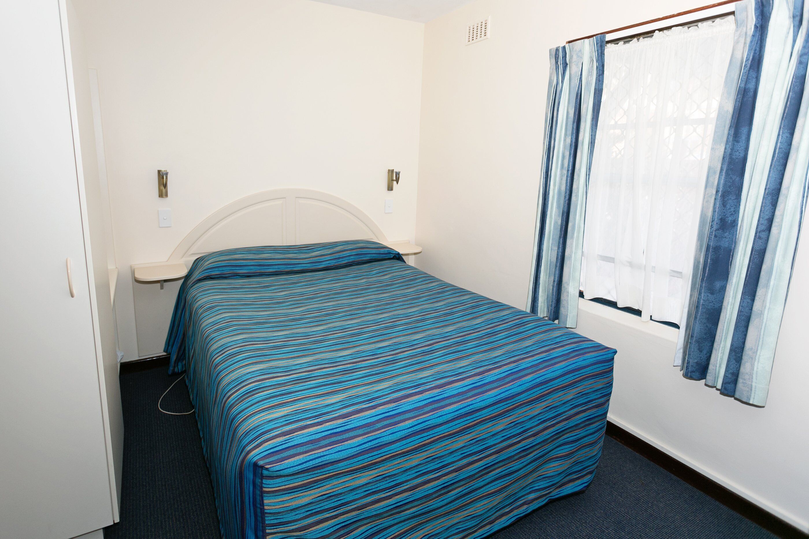 Ein kleines Zimmer mit einem Einzelbett, einer gestreiften Tagesdecke, einem weißen Kopfteil, einem Nachttisch und einem Fenster mit Vorhängen.