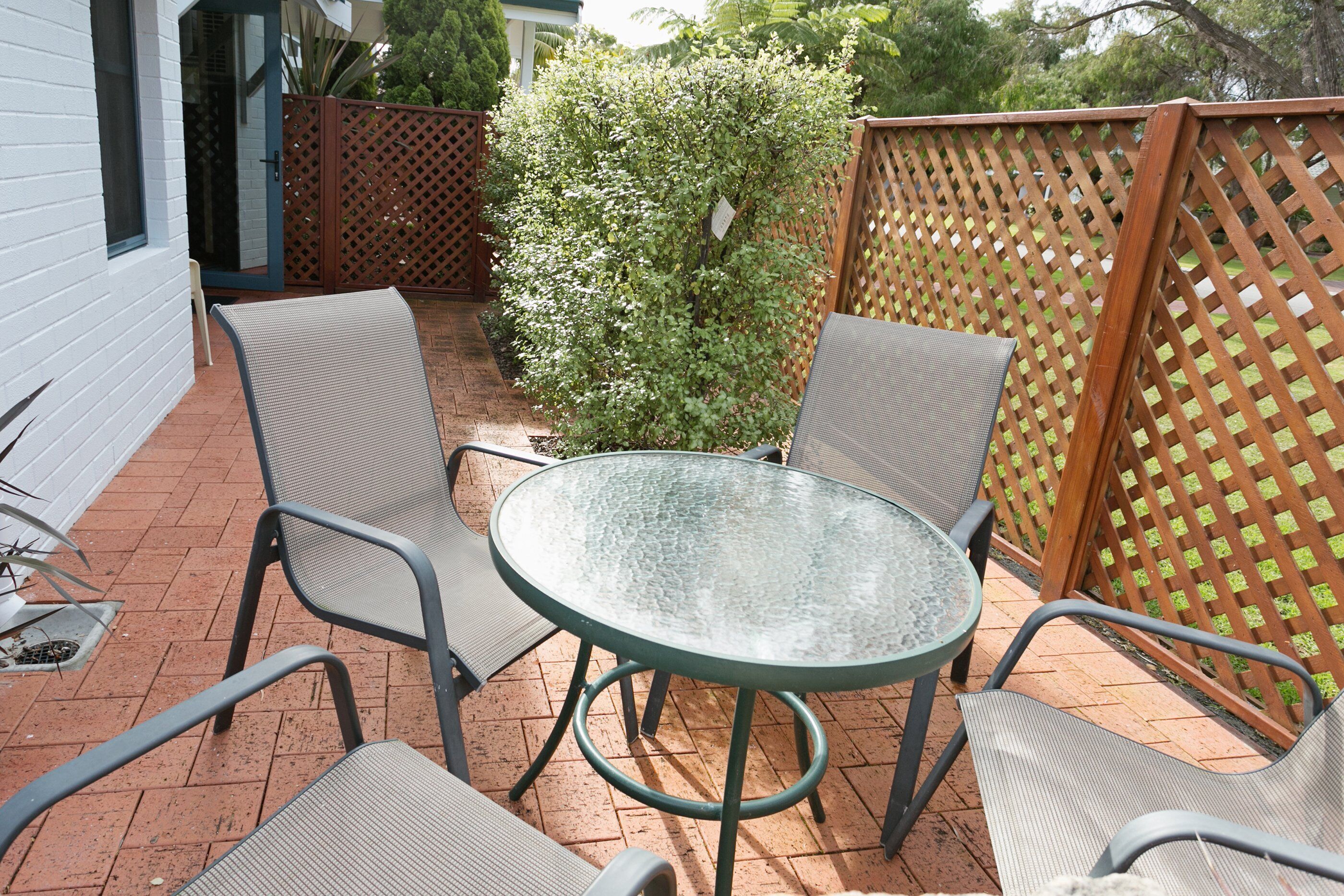 Une terrasse avec une table ronde en verre et quatre chaises, entourée d’une clôture en bois et d’un petit arbre.