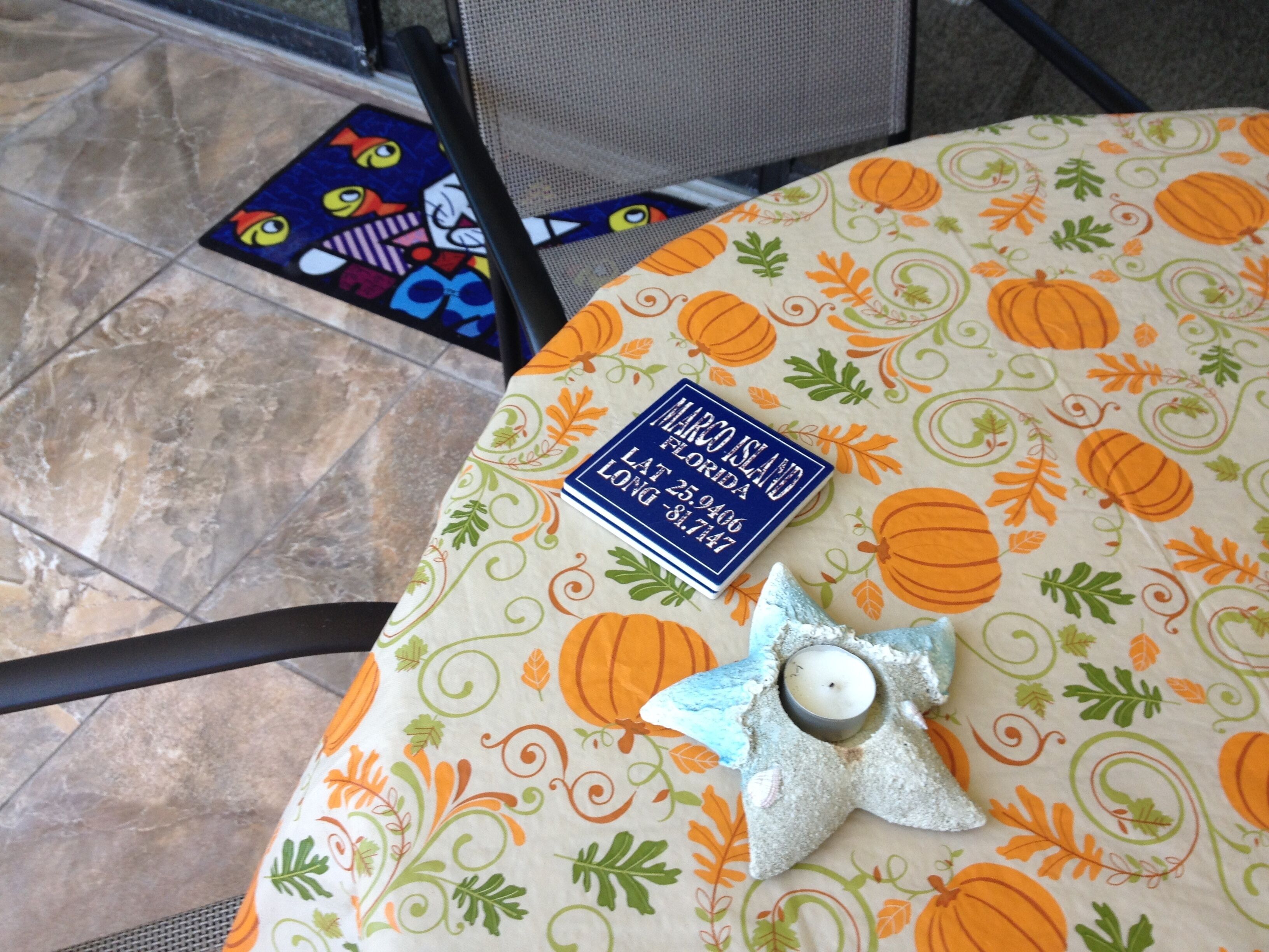 Una mesa con un mantel individual con motivos de calabaza y hojas, una placa con la inscripción 'Marco Island Florida' y un número de teléfono, y un candelabro con forma de estrella con una vela encendida.