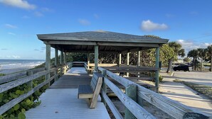A wooden boardwalk leading to a covered pavilion with benches, overlooking a beach.