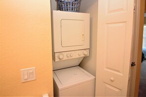 A laundry room with a stacked washer and dryer, a basket on top of the dryer, and a door leading to another room. - Cute and Cozy Venetian Bay Townhome - VB007 (Kissimmee)
