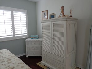 A bedroom with a bed, a white chest of drawers, a small white dresser, a window with blinds, and a framed picture on the dresser. - Perfect Vacation Resort - Private Beach, Golf, Tennis, Pool and More (Hutchinson Island)