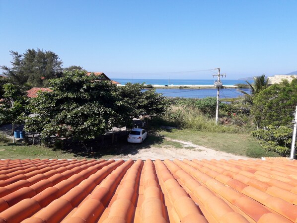 A residential area with a white car parked under a tree, a clear view of the sea, and a tiled roof in the foreground.