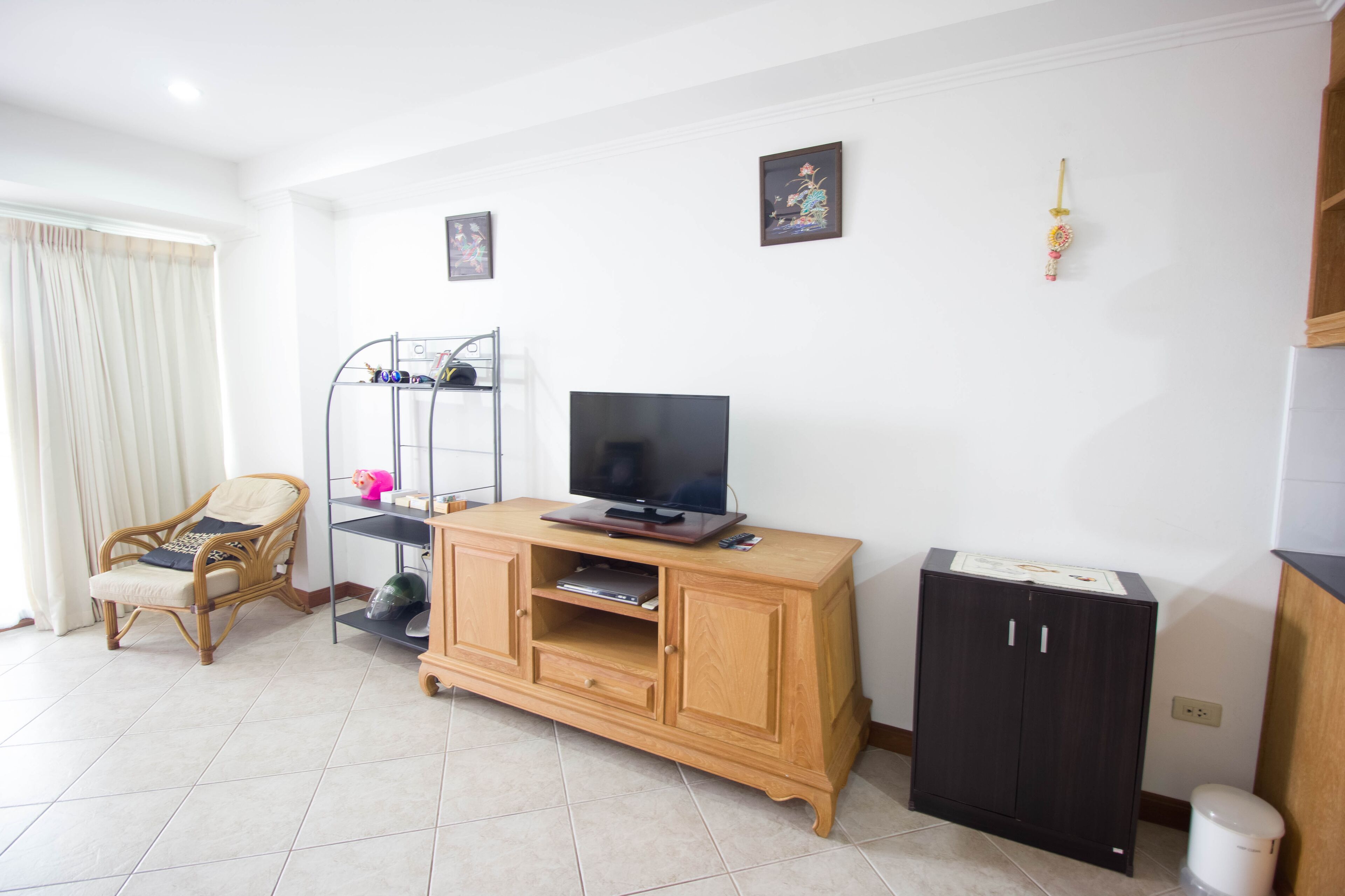 Un salon avec un téléviseur posé sur un meuble en bois, un fauteuil en osier, une petite étagère et un autre meuble orné d’un tableau accroché au mur.