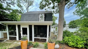 Une maison avec un porche abrité, un grand arbre et deux plantes en pot.