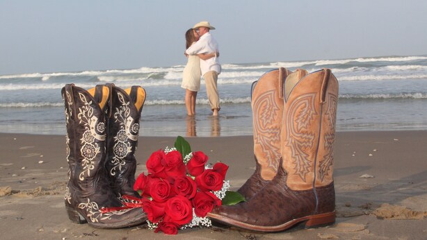 Botas vaquera y un ramo de rosas rojas en una playa con olas al fondo.